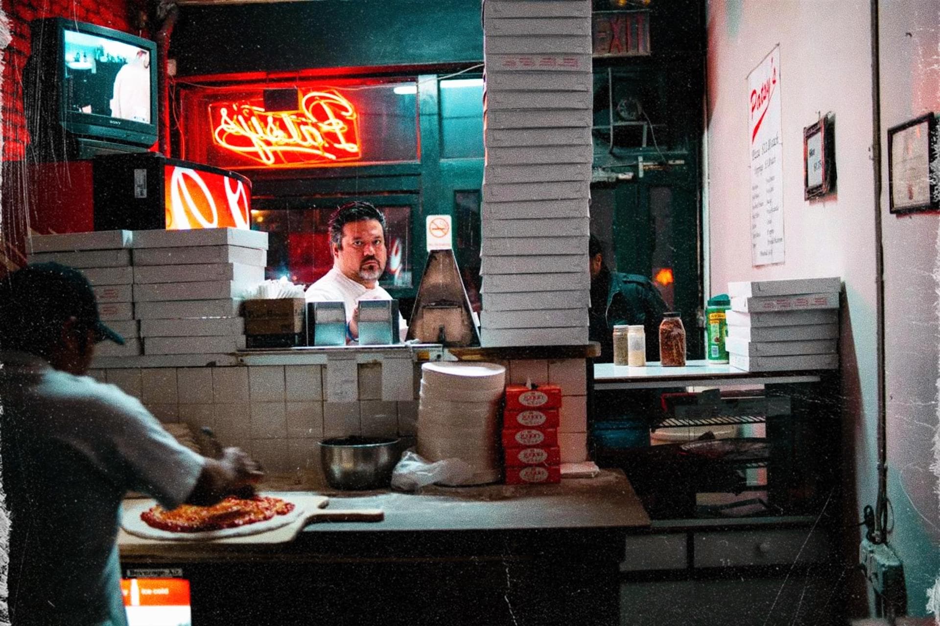 Night inside a New York pizzeria: a worker behind the counter under a red neon sign, stacks of pizza boxes, a pie being made in the foreground.